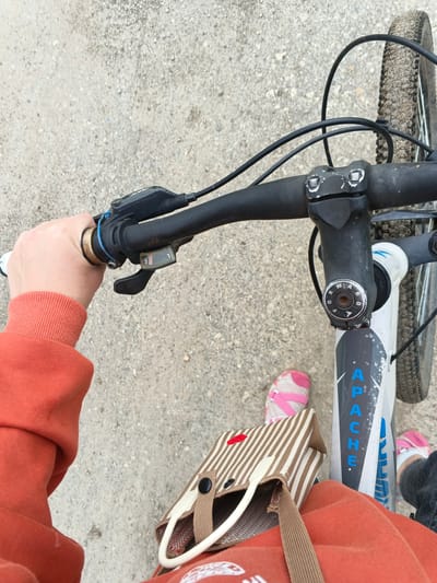 The image shows a first-person perspective of someone riding a bicycle on a gravel path. The rider's left hand, wearing an orange hoodie, is on the handlebars, which are black and have a gear shifter with the number "3" visible. A small brown and white striped bag with red heart detail is hanging from the handlebars. The rider's pink and white sneakers are visible near the front wheel, which is slightly muddy. The bicycle frame is white with blue lettering that appears to spell "APACHE". The background is a grainy, light-colored gravel surface. The location is identified as Medvedevka, Russia.