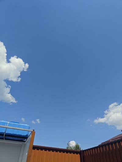 The image depicts a clear blue sky with scattered white clouds, observed from a low angle. In the foreground, a bright blue plastic tank is mounted on a metallic rack atop a white structure. To the right of this structure, an orange corrugated metal fence runs horizontally across the lower portion of the frame, and a darker brown roofline is visible above it. A thin electrical wire stretches across the sky in the mid-ground. The setting appears to be an outdoor area, possibly a backyard or industrial space, in the village of Shkurupiivka, Ukraine, under a sunny sky.
