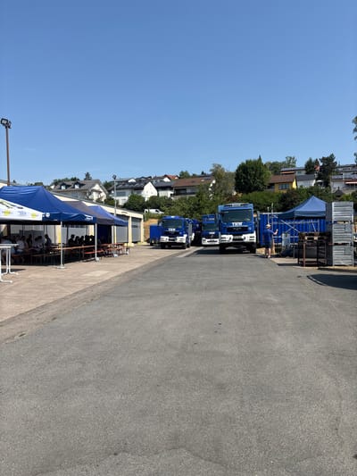 This is a photograph of the Technisches Hilfswerk (THW) facility in Obernburg am Main, Germany, on a clear, sunny day. In the foreground, a paved area leads towards several blue tents and a building with garage doors. People are seated at tables under the tents, appearing to be engaged in a gathering or meal. Further back, three blue THW trucks are parked in a line. One truck has a person standing near it, facing away from the camera. In the background, residential buildings with tiled roofs are visible on a hillside, interspersed with trees. The overall scene suggests an operational facility with a social event or gathering taking place.