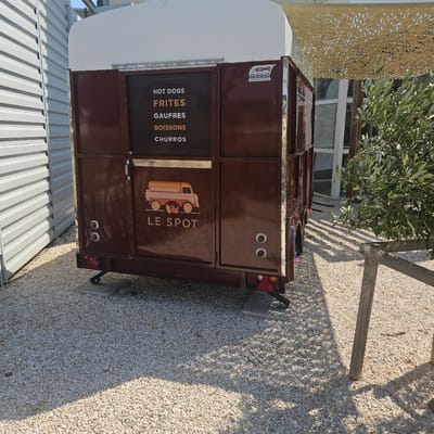 The image shows a dark brown food trailer parked on a gravel surface. The trailer has a white roof and a dark brown front with various food items listed in orange text: "HOT DOGS," "FRITES," "GAUFRES," "BOISSONS," and "CHURROS." Below the text is an illustration of a food truck and the words "LE SPOT." A sticker on the right side of the trailer reads "HEDIMAG FABRICANT DE COMBIS FOOD TRUCK." The trailer is located in La Palme, France, as indicated by context from other images in this dataset which show the same food trailer in the same location. The environment appears to be an outdoor area with a corrugated metal building to the left and a tree with green leaves and a wooden fence in the background to the right. The ground is covered in small, light-colored gravel.