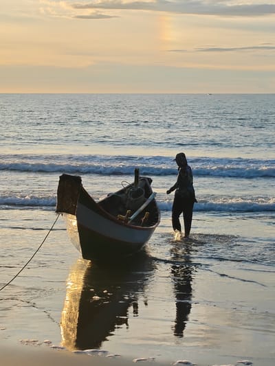 In Uleekareung, Indonesia, a lone individual clad in a dark head covering and pants walks through shallow water at the edge of a beach. The person is moving away from a small fishing boat moored on the wet sand, with waves gently lapping at its hull. The boat, which appears to be made of wood with a colorful stripe along its side, has a patterned cloth draped over its bow. The setting sun casts a warm, golden glow across the scene, reflecting off the water and illuminating the wet sand. The sky above is a soft gradient of yellow and orange, with a few scattered clouds, and a faint rainbow-like atmospheric phenomenon is visible on the horizon. The ocean stretches out, with small waves rolling towards the shore.