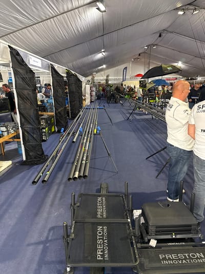 An indoor exhibition, identified as being in Mora, Portugal, is housed within a large tent-like structure with a white fabric ceiling, visible support framework, and overhead rectangular lighting fixtures. The floor is covered with blue carpet. In the immediate foreground, two black "PRESTON INNOVATIONS" branded fishing trolleys are positioned; the one on the right also features a "VENTALITE" label and has a black seat box or tackle box attached. To the left, a collection of approximately ten long, multi-section fishing rods or pole segments are laid horizontally on the carpet, with some displayed on black tripods. Further left, three tall, black, mesh-like bags or storage sleeves hang vertically from a display stand. In the mid-ground, a "LOWRANCE" banner is visible, and numerous other fishing equipment displays are set up, including various rod rests, stands, and two open umbrellas (one black, one blue). Approximately 10-15 individuals, primarily men, are scattered throughout the exhibition space, observing the displays. In the right foreground, two men are visible from the waist up. The man on the left is bald, wearing a white short-sleeved polo shirt with "SPORTING CLUB PORTUGAL" branding and blue jeans. The second man, partially obscured, wears a white shirt with "SPORTING CLUB" text visible and grey jeans. Both appear to be standing and observing the area.