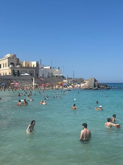 This image depicts a densely populated coastal bathing area in Nardò, Italy, characterized by a clear blue sky and calm, turquoise-blue water.

Approximately 150-200 individuals are present within the scene, primarily adults and some children. A significant concentration of people is seen wading, swimming, or standing in the clear water, with some using flotation devices. Others are situated on the narrow, sand and pebble beach visible in the mid-ground, many gathered under numerous multi-colored beach umbrellas (predominantly red and blue).

The background features a prominent, light-colored stone retaining wall or promenade running parallel to the coastline, equipped with multiple sets of stairs providing access to the beach. Above this structure, several multi-story buildings, consistent with Mediterranean architectural styles, display light-colored facades, arches, and balconies, lining an elevated area. Streetlights are positioned along this elevated section. To the far right, natural rock formations extend into the sea, forming part of the cove's edge. A few yellow recreational kayaks are discernible on the beach near these rocks. The scene is illuminated by bright daylight, suggesting midday or early afternoon.