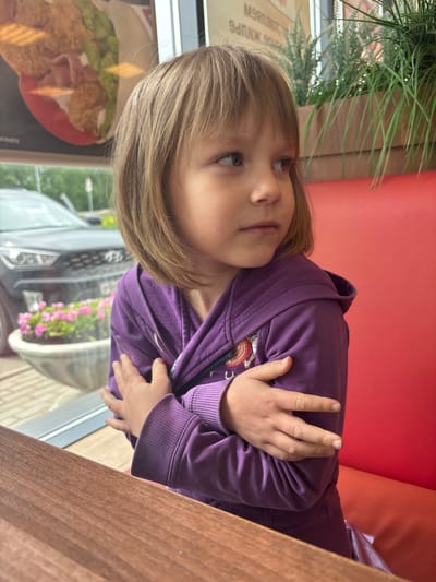 A young child, appearing female, with light brown, bob-cut hair, is seated at a wooden table in an establishment in Krilovo, Russia. She wears a purple zip-up hoodie, with her arms crossed over her chest, hands gripping her upper arms. Her gaze is directed towards the upper right, off-frame. To her right and behind her, a red upholstered booth backrest is visible. Behind the child, through a large window, a dark-colored Hyundai SUV is visible on a street, alongside a white outdoor planter containing pink petunia-like flowers. Inside, behind the child, artificial green plants are arranged in planters, and a menu board displaying inverted Cyrillic text, partially legible as 'МЯСО. БУРГЕРЫ. НАПИТКИ.' (Meat. Burgers. Drinks.), is visible. The outdoor lighting suggests an overcast sky.
