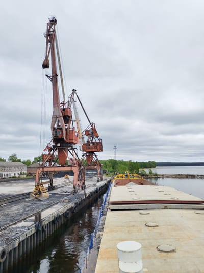 The image depicts an industrial port scene in Medvezhyegorsk, Russia, under an overcast sky, likely on Lake Onega. The central subjects are two large, aged, orange-red industrial gantry or portal cranes positioned along a concrete quay. The crane on the left features a substantial open clamshell grab bucket, and "52" is visible on its cabin. The crane on the right also displays "43" on its cabin. Both cranes show considerable rust and wear.

Piles of dark aggregate, resembling coal or gravel, are scattered across the dock, with a smaller, lighter pile of sand or gravel further back, where a yellow excavator is operating. The quay itself is concrete, reinforced with vertical pilings and worn rubber fenders, reflected in the dark water alongside.

A long, flat-decked barge or cargo ship is moored on the right, its deck covered with numerous round hatch covers. The yellow bow section of the vessel appears to be loaded with bulk material. In the background, a forested shoreline stretches along the lake under the cloudy sky, revealing a sandy beach area. Two small, distant figures are visible on the far shore. On the left, several old, multi-story industrial buildings with multiple windows complete the utilitarian setting. The scene suggests ongoing cargo handling operations in a well-established, though aged, industrial port.
