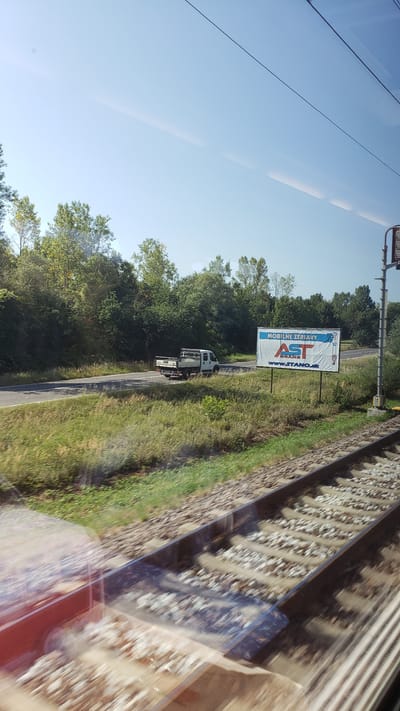 The image captures a view from a moving train in Kostolná-Záriečie, Slovakia. The foreground shows a close-up of train tracks, with a blurred red object on the left side, possibly part of the train carriage. Beyond the tracks, a grassy embankment leads to a road where a white flatbed truck is driving. To the right of the road, a billboard displays an advertisement for "AST MOBILNÉ ŽERIAVY" with the website "WWW.STANO.SK". The background consists of lush green trees and a clear blue sky. The overall scene suggests a rural or semi-rural setting on a sunny day.