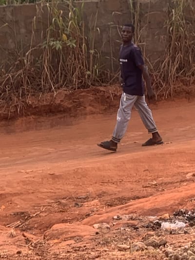 A person stands on a dirt road. The individual wears a dark shirt, light gray pants, and dark sandals. A cap or head covering is also visible. The surrounding environment consists of dry vegetation along the left side of the road, and a mix of vegetation and what appears to be debris on the right side. The earth is a reddish-brown color, indicative of the soil.