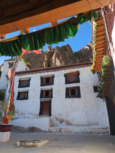 This image shows a courtyard in front of a white-washed building with red-trimmed windows and doors. Prayer flags are strung across the courtyard and hang from a wooden structure overhead, adding a splash of color to the scene. In the background, rocky mountains rise against a clear blue sky, suggesting a remote, high-altitude location. The building itself appears to be old, with some signs of wear and tear on its facade. The courtyard is paved with concrete, and a small, stone platform sits in the foreground. The overall impression is one of a peaceful, spiritual place, likely a monastery or temple, nestled within a dramatic natural landscape.
