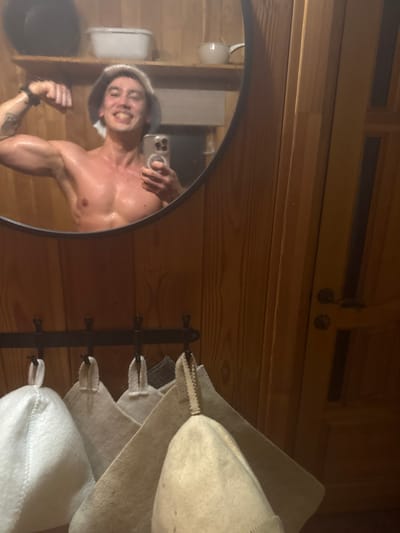 Inside a wooden sauna, a muscular man with blonde hair takes a selfie in a circular mirror. He flexes his left bicep, showcasing a tattoo. He holds an iPhone in his right hand, capturing his reflection. The sauna's wooden paneling is visible, along with shelves, a sink, and a black rack displaying several felt sauna hats. The hats are various shades of white and gray.