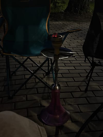 An outdoor scene at night shows a hookah setup on a brick-paved surface. A teal folding chair is positioned to the left, with a dark object, possibly another chair, to the right. The hookah is made of metal with a glass base filled with purple liquid. A red glowing coal rests on top of the hookah bowl, indicating it is in use. The ground is paved with rectangular bricks. A large tree is visible in the background, with foliage and darkness suggesting it is nighttime. The location is specified as Doma 1096 Km, Russia.