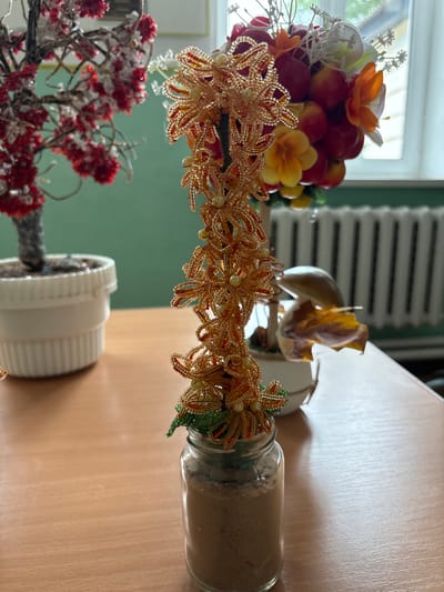 An indoor still life is depicted on a wooden desk, suggesting a location in the city of Ветрынскі сельскі Савет, Belarus. The primary subject is a beaded sculpture of a plant, fashioned with golden and red-orange beads to resemble flowers and green beads for leaves. This sculpture is housed in a clear glass jar filled with a light brown granular substance, possibly sand or small pebbles, serving as its base.
To the left of the beaded sculpture, a potted plant with red bead-like flowers and white accents is visible in a white pot. In the background, a bouquet of artificial fruits and flowers, including red berries and orange and yellow blossoms, is arranged. Behind the bouquet and to the right of the beaded plant, a mushroom-shaped object with a brown and yellow leaf resting on it is partially visible, further contributing to the decorative, possibly autumnal, theme.
The environment includes a muted green wall in the background, with a radiator visible along the lower portion of the wall. A window with light streaming through it is also present in the upper right corner of the frame. The lighting appears to be natural, coming from the window, and casts soft shadows on the desk. There are no people or animals in the image. The overall scene conveys a sense of indoor decoration or craft display.