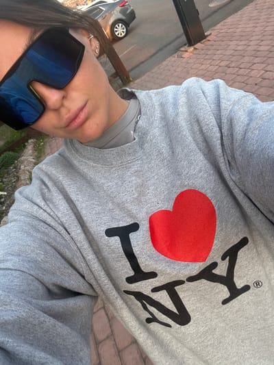A woman wearing oversized sunglasses is taking a selfie outside. She is wearing a gray sweatshirt with a red heart and black lettering. Part of a car and brick walkway are visible in the background. The location is Кузнецово, Russia.