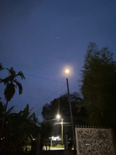 The image depicts a dusky scene in Meunasahlhok, Indonesia. The sky is a deep blue, transitioning into twilight. A bright, round moon is prominently visible in the upper left quadrant, casting a soft glow. To the left of the moon, a faint, elongated streak of light, possibly a distant aircraft or celestial body, is present.
In the foreground, silhouetted against the sky, are the fronds of palm trees, suggesting a tropical environment. A wooden utility pole with wires stretches across the frame, with a streetlamp near its base illuminating a small area. To the right, a decorative, white metal gate with an ornate, swirling pattern is visible, suggesting a residential or property boundary. Beyond the gate, lights from what appears to be a building or a street are discernible, indicating the presence of human activity. The overall mood is tranquil and serene, typical of an evening setting. The time of day is clearly dusk or early night. No visible text is present in the image.