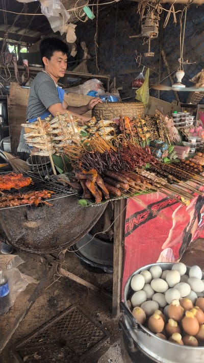 A middle-aged Asian man is standing behind a food stall in Phao, Laos, arranging skewers of grilled food. He wears a grey t-shirt and a blue apron. The stall is laden with various grilled items, including fish on skewers, meat skewers, and what appear to be chicken wings. A large, dark, cauldron-like pot is visible to the left, likely used for grilling. In the foreground, a metal pot is filled with boiled eggs, some of which are peeled, revealing a bright yellow yolk. The stall is constructed from rough materials, with a corrugated metal roof and bamboo fencing. The ground is dirt and appears somewhat muddy, suggesting an outdoor market setting. Plastic bags and other miscellaneous items are hanging from the stall's structure. A red and white banner or sign is draped across the front of the stall. The overall impression is of a busy, rustic food vendor operating in a local market.
