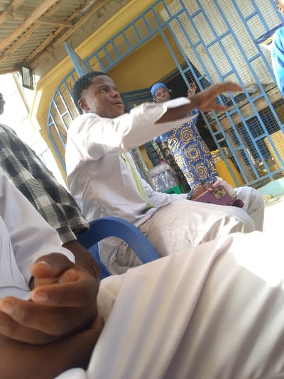 A man seated, gesturing with his right hand, wears a white garment with lime green trim. He sits on a blue chair, phone in lap. A woman stands behind him, wearing a blue and brown patterned dress and a blue headscarf. Another man, partially visible, wears a white garment with a gray and black plaid shawl. A blue metal gate is behind them. Bottles of water are visible in the background. The perspective is angled upward.