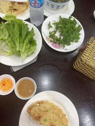 A table is set with various dishes: a plate of fresh herbs, a plate of lettuce, and a crispy pancake-like item. There are also small bowls of dipping sauces, one with a tan color and another with a red-tinged sauce. A water bottle lies next to the greens. A woven container sits at the top of the frame, and a curved dark stain mars the table surface.