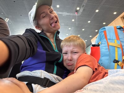 A close-up, low-angle selfie shows a woman wearing a light green baseball cap and a black and blue jacket, with her tongue sticking out and eyes closed in a playful expression. She is holding a young boy who is crying and looking directly at the camera. The boy is wearing an orange t-shirt. In the background, a blue and orange backpack is visible, along with a red object. The ceiling appears to be a metallic grid with overhead lighting. The scene is indoors, possibly at an airport or public waiting area. The location is not specified as the city of Bolshiye Kabany, Russia.