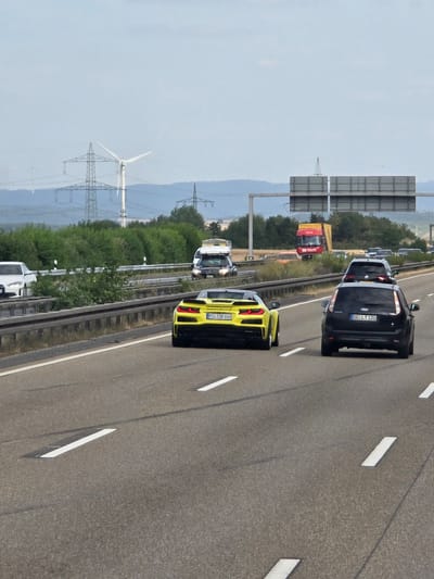 An overcast day on a German autobahn, likely the A3 near Limburg an der Lahn, Germany, as indicated by the presence of typical German road signs and the landscape. In the foreground, a bright yellow Chevrolet Corvette Stingray is in the right lane, its rear end facing the camera. To its right and slightly ahead, a black Ford Focus is also in the right lane. In the left lane, a white BMW sedan is visible in the distance, traveling in the same direction.

Further down the autobahn, a red and yellow truck is in the right lane, followed by several other vehicles. Overhead, large gray signs with white lettering indicate upcoming route information. The landscape beyond the autobahn consists of rolling hills with patches of trees and fields. In the far distance, several wind turbines are visible, along with large electricity pylons. The road surface is asphalt, with clear white lane markings. The overall scene depicts a typical highway traffic situation in Germany.