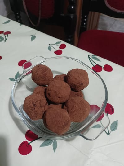 A close-up, overhead shot shows a glass bowl filled with chocolate truffles. The truffles are dark brown and dusted with cocoa powder, with a somewhat irregular, spherical shape. They are arranged in the clear glass bowl, which has a fluted rim. The bowl rests on a white tablecloth patterned with red cherries and green leaves. In the background, the dark wood of a chair is visible, along with a sliver of a red chair cushion. The lighting appears to be natural, casting soft shadows. The scene is likely from a home setting.