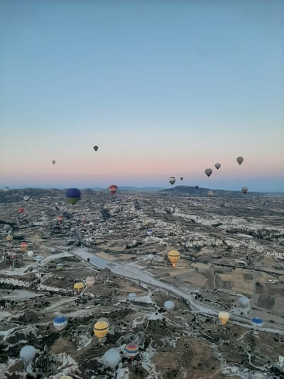 An aerial, wide-angle shot captures a panoramic view of a valley filled with hot air balloons, set against a soft twilight sky. The landscape is rugged and mountainous, with a winding river or road running through the valley floor. The terrain is a mix of barren, rocky outcrops and cultivated fields. Dozens of hot air balloons are scattered across the sky, varying in size, color, and pattern, with some displaying brand logos. The balloons appear to be ascending or drifting leisurely. The scene depicts a popular tourist activity, likely a sunrise balloon ride over the unique geological formations of Cappadocia, specifically the region near Avanos, Turkey, known for its fairy chimneys and dramatic landscapes. The overall atmosphere is serene and breathtaking, highlighting the beauty of the natural environment and the spectacle of the balloons.
