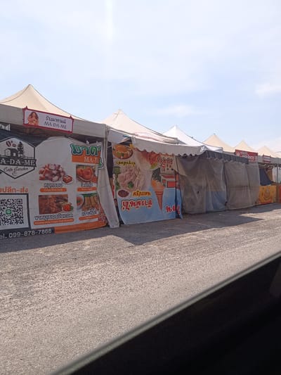 The image captures a row of food stalls under white tent-like structures along a paved road, possibly within Hua Hin City Municipality, Thailand. Each stall is marked with advertising signage displaying various Thai dishes, including crispy pork belly, shrimp donuts, German-style crispy pork knuckle, and spring rolls. The first stall on the left has a prominent sign labeled "A-DA-ME Cafe Garden" with a QR code and a phone number. Another stall features a graphic of an ice cream cone and a text reading “ข้าวมันไก่”. The clear blue sky suggests it's daytime. The image appears to be taken from inside a vehicle, indicated by the car door in the foreground.