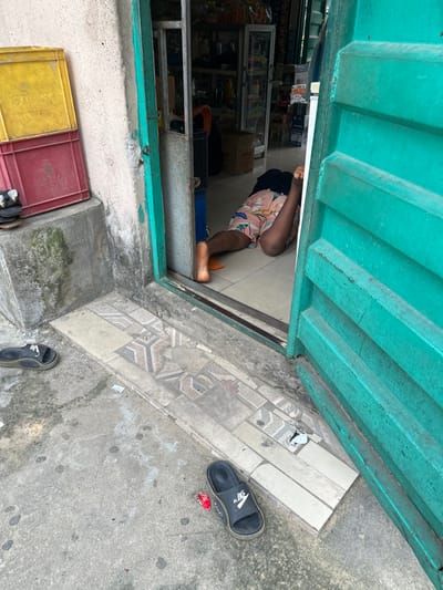 From an overhead perspective, the image captures a person partially inside a shop. They are bent over, with their head and torso inside and feet and legs outside. The shop is filled with shelves of goods. Outside the shop, there is a green door, a tiled entrance, and a concrete ground. Scattered on the ground are sandals, a red object, and yellow and red crates.