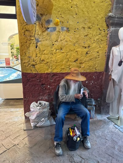 An eye-level, indoor shot, shows an elderly man, dressed in a hat, long beard, gray jacket, jeans and athletic shoes, sitting and reading while holding a small book in his hands. The man sits on a black chair, partially obscured by his legs and body. To his right, a black backpack rests on the ground, filled with various objects including a yellow and red bag, and a bag of chips. Next to the backpack is a full, partially crumpled sack.
The man sits in a corner of the store, surrounded by walls of yellow and maroon. Behind him is a statue. To the left, a clock hangs on the wall. In the foreground is a display of goods for sale. The floor is brick, laid in a pattern.
The scene is the city of San Miguel de Allende, Mexico.