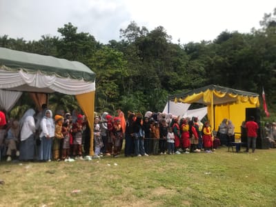 In Pantonrayeuk Satu, Indonesia, an outdoor event is taking place under a cloudy sky. Two large tents, one green with white drapes and the other yellow with white drapes, are set up on a grassy field. A line of people, including children and adults, are standing facing away from the camera, seemingly watching a performance or ceremony. Many of the individuals are wearing traditional attire, including headscarves and colorful garments. Some children are dressed in matching outfits, suggesting a group performance. In the background, a dense forest covers a hillside. To the right of the yellow tent, a black speaker system is visible, and a man in a red shirt is standing near it. The overall scene suggests a community gathering or cultural event.