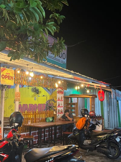 This is a vertical, nighttime photograph of an outdoor establishment in Boja, Indonesia. The scene depicts a street food or casual dining area, likely a warung or restaurant, with several motorcycles parked outside. The building itself has a brightly lit yellow exterior with red and green accents.

Prominently featured are several large, colorful banners. One banner displays the text "MARMIN" in large, stylized letters, possibly indicating the name of the establishment. Another sign reads "OPEN" in bold red and yellow lettering. Signs with Asian script, specifically Japanese characters, are visible, including on a red lantern hanging from the awning. There are also banners with images of food items.

A few people are present. One man is seated at a table, looking at his phone. Another individual, wearing a helmet, is partially visible near a motorcycle. A person in an orange jacket is also near the motorcycles.

The setting is illuminated by a string of lights overhead and the lights from the building itself, creating a warm and inviting atmosphere. The presence of motorcycles suggests this is a popular spot for locals to gather. The overall impression is that of a lively, casual dining experience at night.
