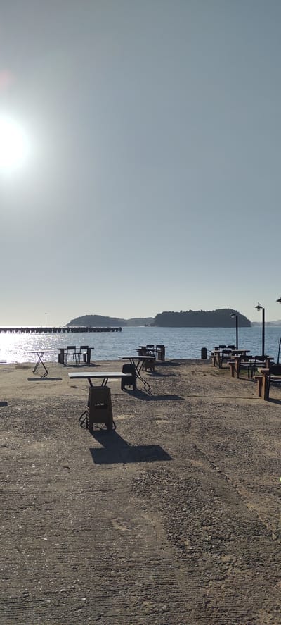 The image shows an outdoor seating area near a body of water, likely the sea, under a clear, bright sky with the sun prominently featured in the upper left quadrant. The foreground is a concrete ground surface with scattered gravel and visible tire marks. Several empty tables and chairs are set up, some made of metal with a distressed finish, others appearing to be plastic. The tables are of various designs, including folding tables and sturdier ones supported by stacked plastic crates. Long shadows are cast by the tables and chairs, indicating a sunny day.
In the middle ground, the water stretches out, reflecting the sunlight. A long pier or jetty extends into the water from the left side of the frame. On the right side, there are two street lamps with downward-facing lights. In the distance, across the water, is a hilly, tree-covered island.
There are no people visible in the scene. The overall atmosphere is quiet and undisturbed, suggesting a place that is either closed for the day or not yet open for business. The location in Calang, Indonesia, is not explicitly identified by any signage or landmarks within the image itself, but the description of a coastal setting with islands and a pier aligns with the geographical characteristics of coastal Indonesia.