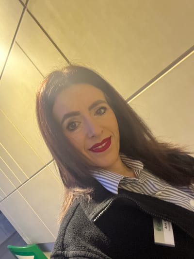A selfie photograph taken indoors. The subject is a woman with dark brown hair, fair skin, and wearing red lipstick. She is wearing a striped shirt under a dark jacket and has a name tag on her jacket labeled "Ana B". The background shows a tiled wall or floor. The image's location is implied to be in Mensdorf, Luxembourg.