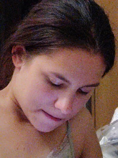 The image shows a close-up of a young person's face, seen from the side, with their head tilted down. Their eyes are partially closed, and their lips are slightly parted. The person has dark hair that is pulled back. The background is textured and appears to be a wall or fabric, with a blurred object on the right side of the frame. The lighting is soft, casting shadows on the face. There is no discernible location in the city of Okhtyrka, Ukraine, visible in this image.