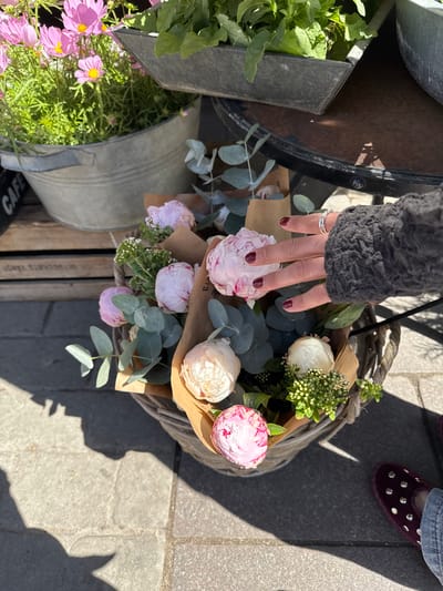 A top-down shot reveals a hand gently touching a bouquet of flowers in a basket. The hand, adorned with a silver ring and polished with dark red nail polish, hovers over pink and white peonies wrapped in brown paper. Eucalyptus leaves are interspersed among the blooms. The basket sits on a wooden crate, beside metal containers filled with greenery and pink daisies. The lower right of the image shows a section of paved ground with a maroon slipper visible near the upper right corner of the frame. The scene suggests an outdoor flower market or shop, possibly located in Malmö, Sweden, given the presence of the Swedish word "BLOMMOR" partially visible on the wooden crate below the basket, a term which means "flowers" in Swedish.