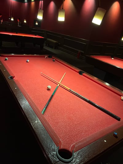 The scene depicts an interior view of a dimly lit billiards hall located in Bayraklı, Turkey. In the foreground, a rectangular pool table with worn, textured red felt dominates the frame. On this table, a white cue ball rests near the center, flanked by two wooden pool cues crossed diagonally. One cue features a dark green handle, while the other has a black handle with a lighter mid-section. Scattered across the red felt are several billiard balls: a striped yellow and white 9-ball, a solid blue 2-ball, a solid green 6-ball, a solid red 3-ball, and a solid brown/orange 1-ball. A small blue cube, possibly pool chalk, lies near the bottom right pocket.
The background reveals a red-walled environment illuminated by multiple pendant light fixtures with rectangular, light-colored shades, casting warm yellow and red glows on the walls. Two additional pool tables, also with red felt, are visible in the mid-ground and far right, with a white ball present on the far right table. Seating, in the form of dark upholstered benches, lines the back wall. A red fire extinguisher is mounted on the wall to the right of the center background. Two indistinct human figures are partially visible in the far-left background, one standing near a pool table. No specific actions or interactions are clearly discernible from these individuals due to distance and focus.