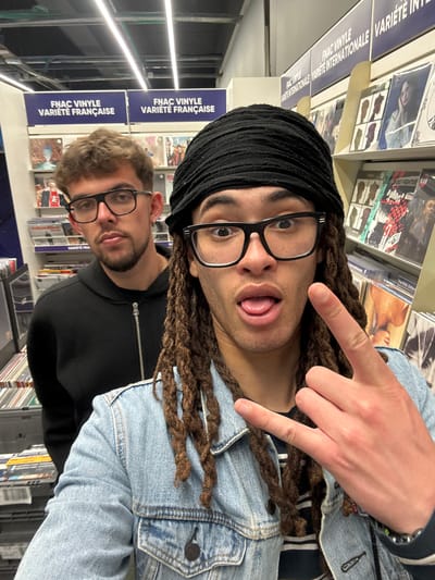 The image shows two individuals in what appears to be the FNAC store in Villebon-sur-Yvette, France. The setting includes racks of vinyl records and related signage. The first person, positioned in the foreground, wears a black head covering, glasses, and a denim jacket. They are sticking their tongue out and making a "peace sign" with one hand. The second person, partially visible behind the first, wears glasses and a black hooded sweatshirt.