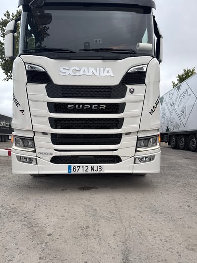 A white Scania 500 S tractor unit is centrally positioned, facing forward, on a light grey gravel surface in San José Obrero, Spain. The truck features "SCANIA" prominently on its grille, with "SUPER" below it, and "500 S" on the lower front bumper. A red Scania Griffin logo is affixed to the grille. The license plate, "6712 NJB," is Spanish, indicated by the blue 'E' on the left. White headlights and lower daytime running lights are illuminated, along with an orange indicator light visible on the right side.

The cab's upper left side displays angled black text "JOSE A.", while the upper right side shows angled red text "MARIO". A large, dark-tinted windshield reflects an overcast sky and sparse tree foliage. A roll of white paper is visible on the dashboard, hanging from the top right interior of the windshield. The truck's side mirrors are white-cased and black.

To the left of the truck, a portion of a dark-colored semi-trailer is visible with the text "upolucas" and "de la huerta a su mesa". Green tree foliage is present behind and above this trailer. To the right of the truck, a white multi-axle semi-trailer is visible, adorned with large grey line drawings of a bull, fish, and plants arranged in a grid pattern. The ground consists of light grey gravel with darker stones and subtle tire tracks, featuring a small dark stain near the front right wheel. A red and white barrier is partially visible to the left of the truck's front wheel. The sky is overcast. No individuals are present in the image.