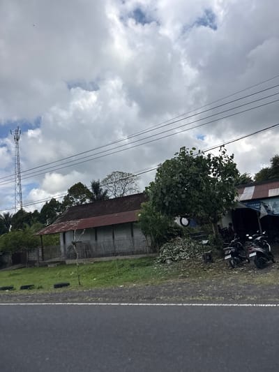 The image depicts a roadside scene in Susut, Indonesia. In the foreground, a grey asphalt road with a white line is visible. Beyond the road, a grassy embankment leads up to a weathered building with a corrugated metal roof. The building appears to be a long, single-story structure with a fence made of vertical wooden slats. To the left of the building, a tall telecommunications tower stands against a cloudy sky. A cluster of trees with dense foliage is situated to the right of the building, partially obscuring another structure with a similar corrugated metal roof, possibly a shop. Two motorcycles are parked in front of this second structure. The overall environment suggests a semi-rural or small town setting.