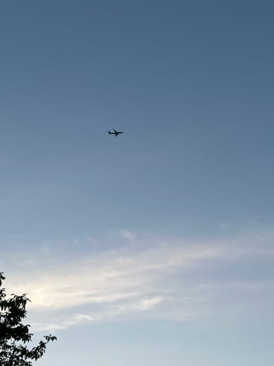 An airplane is flying in the sky over Mondercange, Luxembourg. The airplane is silhouetted against the blue sky. There are some clouds in the sky, and a tree is visible in the foreground.