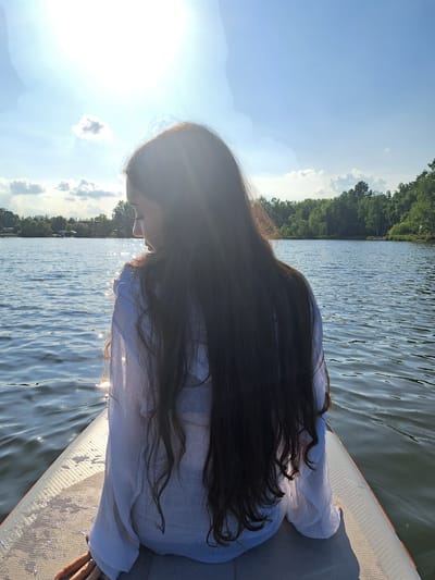 A person is sitting on a paddleboard in the water, facing away from the camera. The person has long, dark hair and is wearing a white shirt. The paddleboard is on a body of water that appears to be a lake or river. The background shows a tree line and a clear blue sky with some clouds. The sun is shining brightly, casting a glare on the water. The overall impression is a serene outdoor scene. The city of Razdolnoye, Russia is not visible in the image.