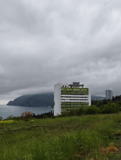 The image depicts a landscape with a grassy field in the foreground. Behind the field, there is a tall, white building with green accents. Further in the background, a body of water extends towards a mountainous landmass shrouded in clouds. To the right of the white building, another cylindrical-shaped white building is visible amongst the trees. The sky is overcast with gray clouds. The scene appears to be in the city of Ut'os, Ukraine.