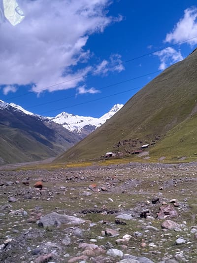 This is a landscape photograph taken from a slightly elevated perspective, looking towards a valley between two imposing mountain slopes. The sky is a vibrant blue, with scattered white cumulus clouds. Power lines are visible stretched across the sky. The left mountain slope is covered in green grass, and in the distance, snow-capped peaks rise dramatically. The right mountain slope is also predominantly green, with a scattering of rocky terrain.
Nestled on the lower part of the right mountain slope, a small cluster of buildings is visible. These appear to be simple structures with dark roofs. Some of the buildings look older, possibly ruins or stone foundations, while others are more intact. The valley floor is a mix of rocky ground, sparse green vegetation, and patches of yellow wildflowers. A few scattered boulders and stones are present in the foreground. The overall impression is one of a remote and rugged mountainous environment, likely in a rural or undeveloped area.
The image does not contain any specific landmarks or features that definitively identify it as being in Almasiani, Georgia. However, the mountainous terrain and vegetation are consistent with landscapes found in Georgia's mountainous regions.
