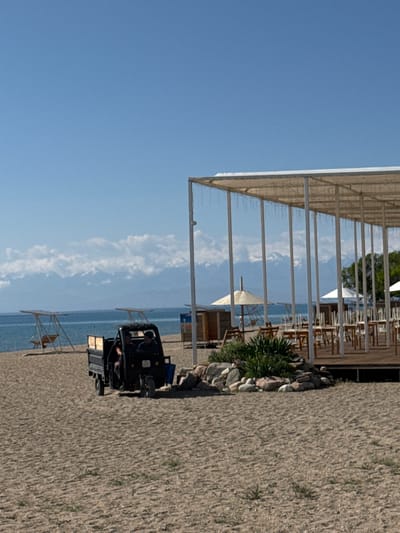 A scene unfolds on a sandy beach in Koshkol', Kyrgyzstan, with a clear blue sky overhead and distant snow-capped mountains forming a backdrop. In the foreground, a three-wheeled utility vehicle is parked on the sand, with two individuals visible inside. To the right, a covered outdoor dining area with white pillars and a white roof is set up, featuring wooden tables and chairs. A few beach umbrellas are visible. In the mid-ground, the calm, blue water of a lake stretches out, and a wooden swing set with a canopy is situated on the beach. The sand is textured with scattered dry grasses.