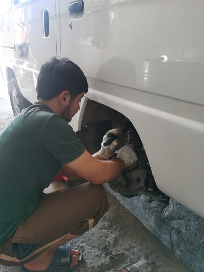 A man is crouched down next to the rear wheel well of a white van, working on the suspension system. He is wearing a dark green polo shirt, brown pants, and white work gloves. The van appears to be in a garage or workshop setting, with a concrete floor visible. The man is holding a tool in his gloved hands, seemingly in the process of adjusting or repairing the shock absorber. The environment suggests a scene of automotive maintenance.