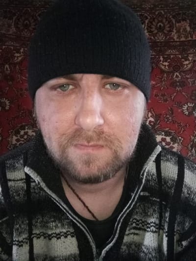 A person with light eyes and stubble, wearing a black beanie and a black and grey plaid zippered sweater, is positioned in front of a patterned red and brown rug. A thin brown necklace hangs below the collar. The location is not evident in the image and cannot be determined.