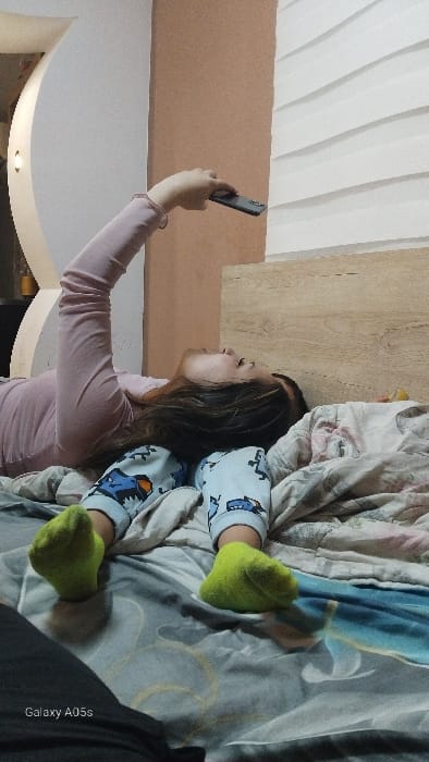 A female individual with long dark hair is depicted lying supine on a bed in the city of Oreshene, Bulgaria. She is wearing a light pink long-sleeved shirt, light blue pajama pants featuring cartoon patterns, and bright lime green socks. Her right arm is extended overhead, holding a dark-colored smartphone, with her head turned slightly to her left, directing her gaze upwards towards the phone. The bed is covered with patterned bedding in shades of blue, grey, and white, with yellow accents. A light wood-grained headboard is visible behind her head. The background wall features a vertical division: an off-white curved architectural element on the left with an embedded warm light fixture, and a light brown/peach-toned wall section on the right, adjacent to a white textured wall with horizontal grooves. The scene is indoors, consistent with a bedroom setting. The lower left corner displays a 'Galaxy A05s' watermark.