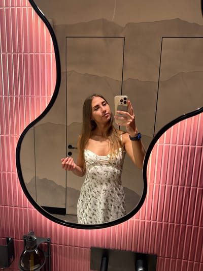 A young woman with long brown hair is taking a selfie in a mirror in a bathroom. She is wearing a white floral print dress and a dark smartwatch on her left wrist. She is holding a smartphone in her right hand, which is angled to capture her reflection. The mirror has an unusual, wavy black frame and is mounted on a wall tiled with vertical pink rectangular tiles. The wall behind the mirror appears to have a subtle abstract pattern. To the left of the mirror, a dark dispenser with a glass bottle is mounted on the tiled wall. The overall scene suggests a modern and stylish bathroom setting.