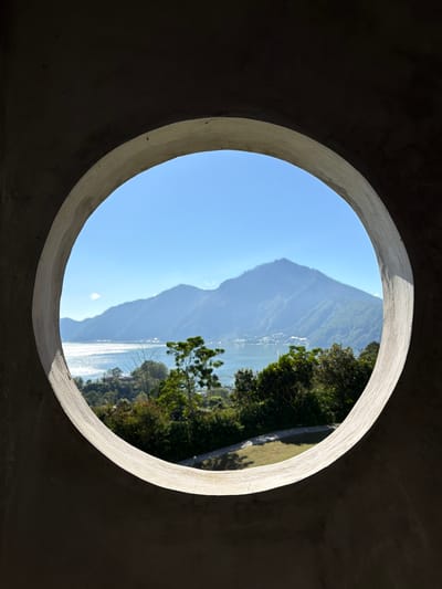 The image depicts a view through a circular concrete portal, framing a landscape in Kedisan, Indonesia. Dominating the scene is a large, hazy mountain rising sharply from the water. The water reflects sunlight. The foreground consists of lush green vegetation along the water's edge. The circular opening is made of a light-colored concrete, casting shadows on its inner surface. The surrounding wall appears to be made of the same concrete. The sky is clear and blue.