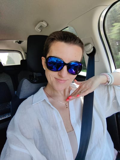 A woman with short, dark hair and sunglasses is sitting in the driver's seat of a car, with her seatbelt fastened. She is wearing a white, unbuttoned shirt over a beige tank top, and a delicate necklace. Her left hand, with bright coral nail polish, is resting on her neck. She is looking at the camera and smiling. The interior of the car, including the headrest and seatbelt, is visible. Outside the car, trees and greenery can be seen through the window, suggesting a rural or suburban setting. The image appears to have been taken in the city of Ilyanskiy selskiy Sovet, Belarus.
