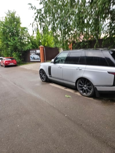 The image shows a silver Range Rover parked on a wet asphalt road. A red car is visible in the background to the left, also parked on the road. Behind the cars, a dark wooden fence runs along a property line, with a signpost that appears to be a "MOTOR TECH" advertisement featuring a white car. Lush green trees with a blurred effect due to motion are present behind the fence, suggesting the image was taken with a moving camera. The overall environment appears to be a residential or commercial street in Sosnovoborsk, Russia, possibly during or after a rain shower given the wet road surface and motion blur.