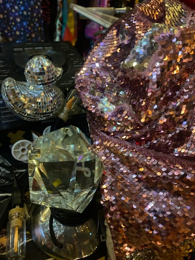 The image shows a close-up of various objects. A section of fabric covered in iridescent sequins is at the top of the frame. Below the sequins is a small mirrored disco ball, a clear geometric prism, and a silver lamp base. A small glass bottle with a filament bulb is also visible. The environment appears cluttered. The location is Baan Nai Suan, Thailand.
